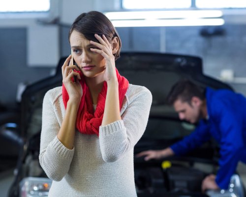 Worried customer talking on mobile phone while mechanic examining car in background at repair garage